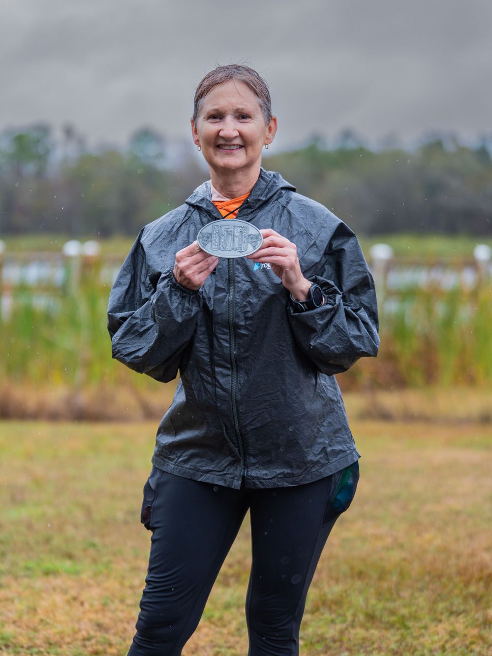 Runner in rain jacket proudly displays finisher medal at outdoor event.