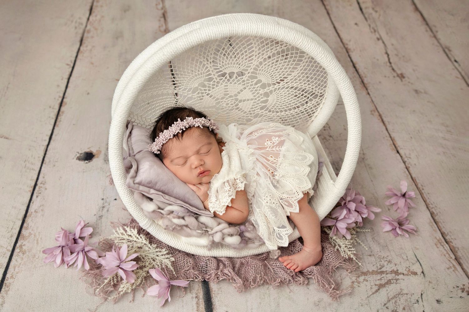 Sleeping newborn in white lace dress posed in round white basket with purple flowers on wooden floor.