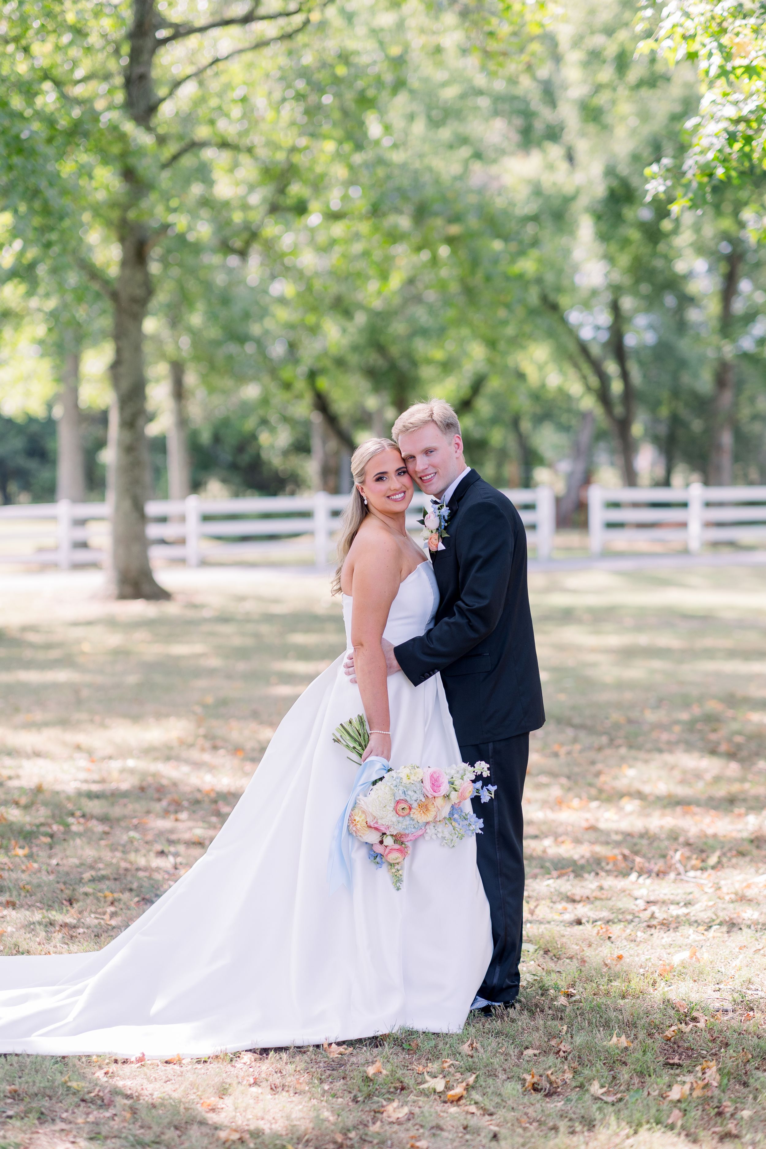 A couple poses for wedding photos in a rustic outdoor setting with white fencing and trees.