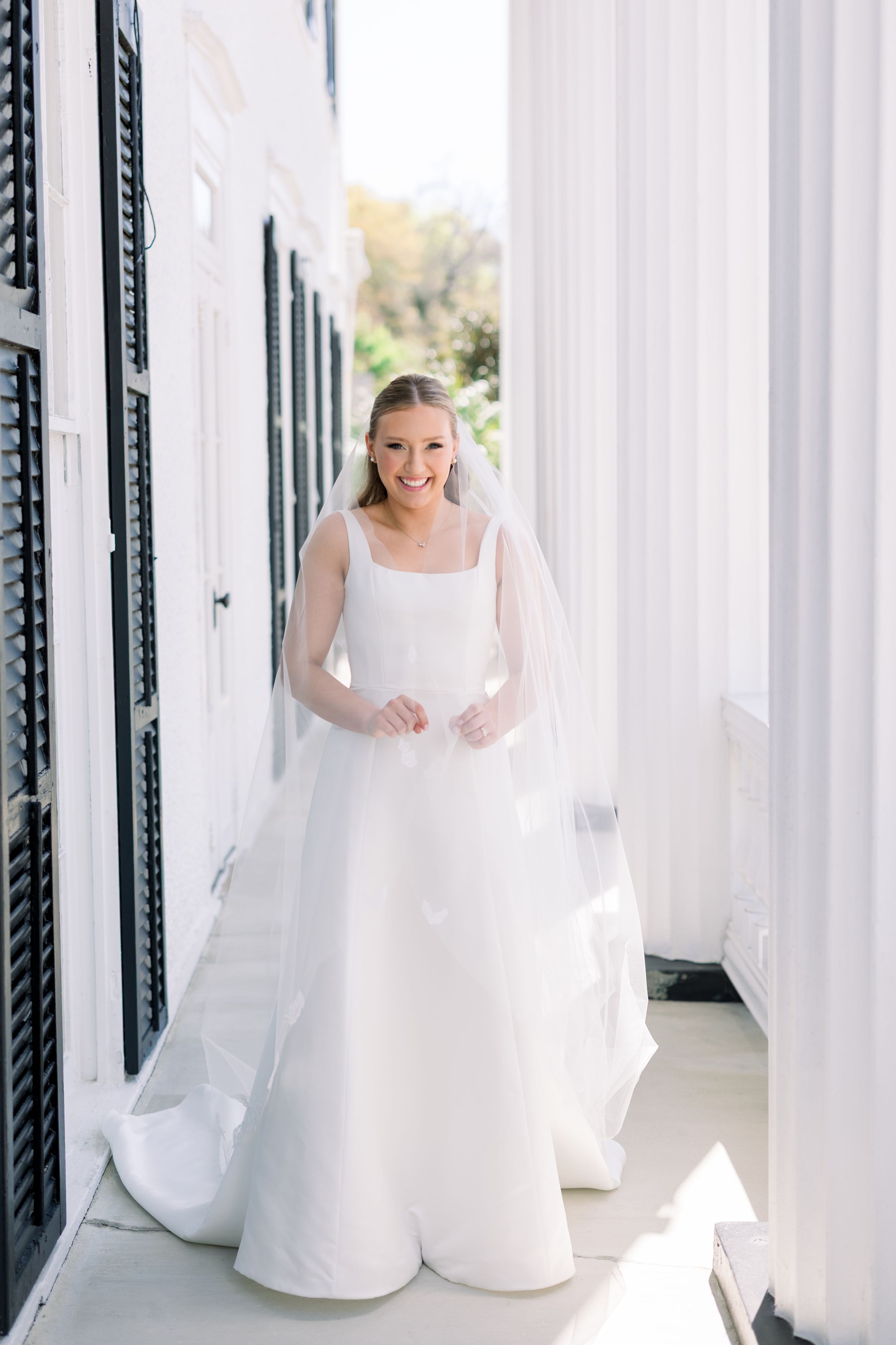 Bride in elegant white gown standing on classic southern porch with white columns.