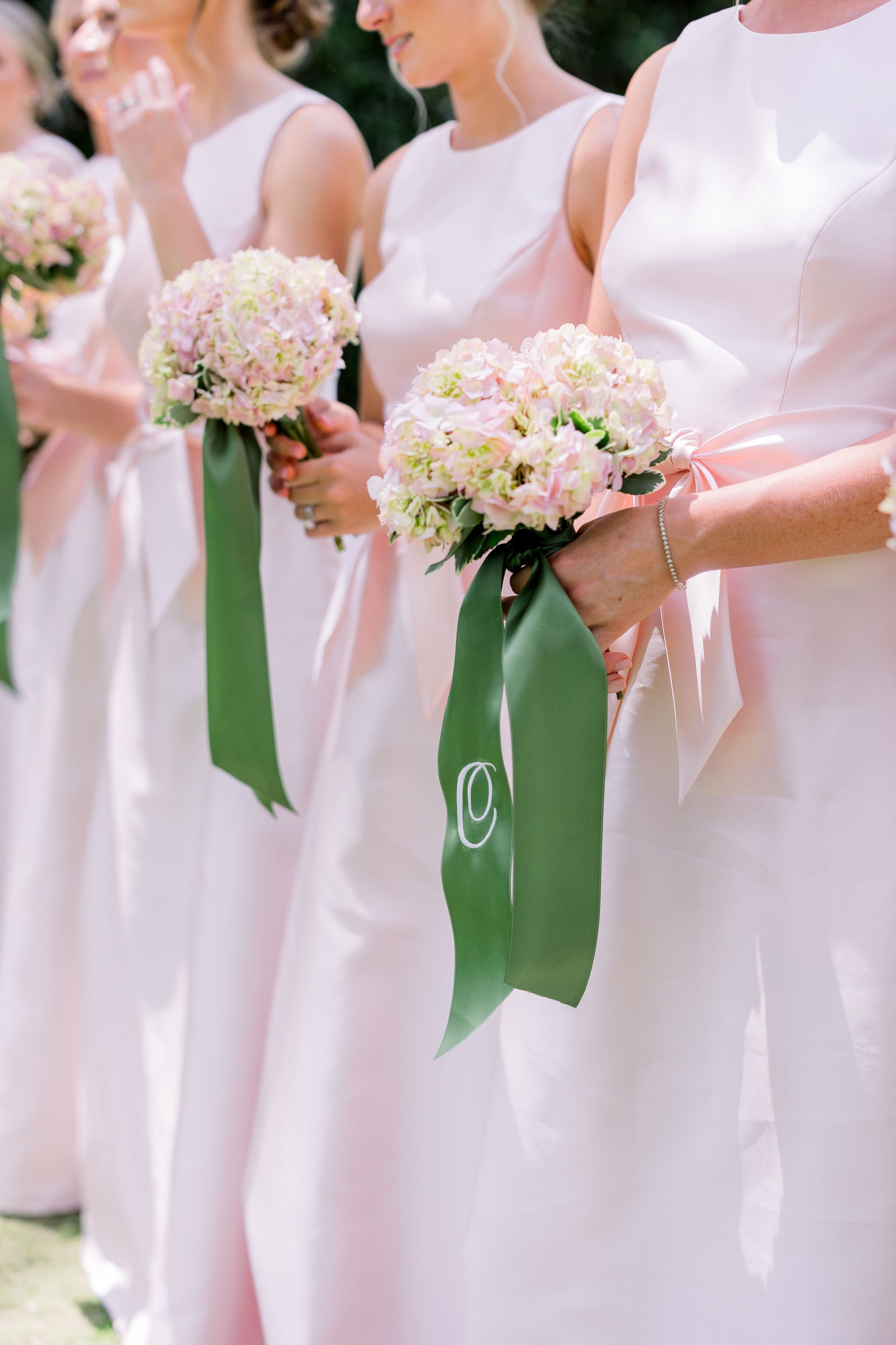Row of bridesmaids in pale pink dresses holding matching bouquets at an outdoor wedding.