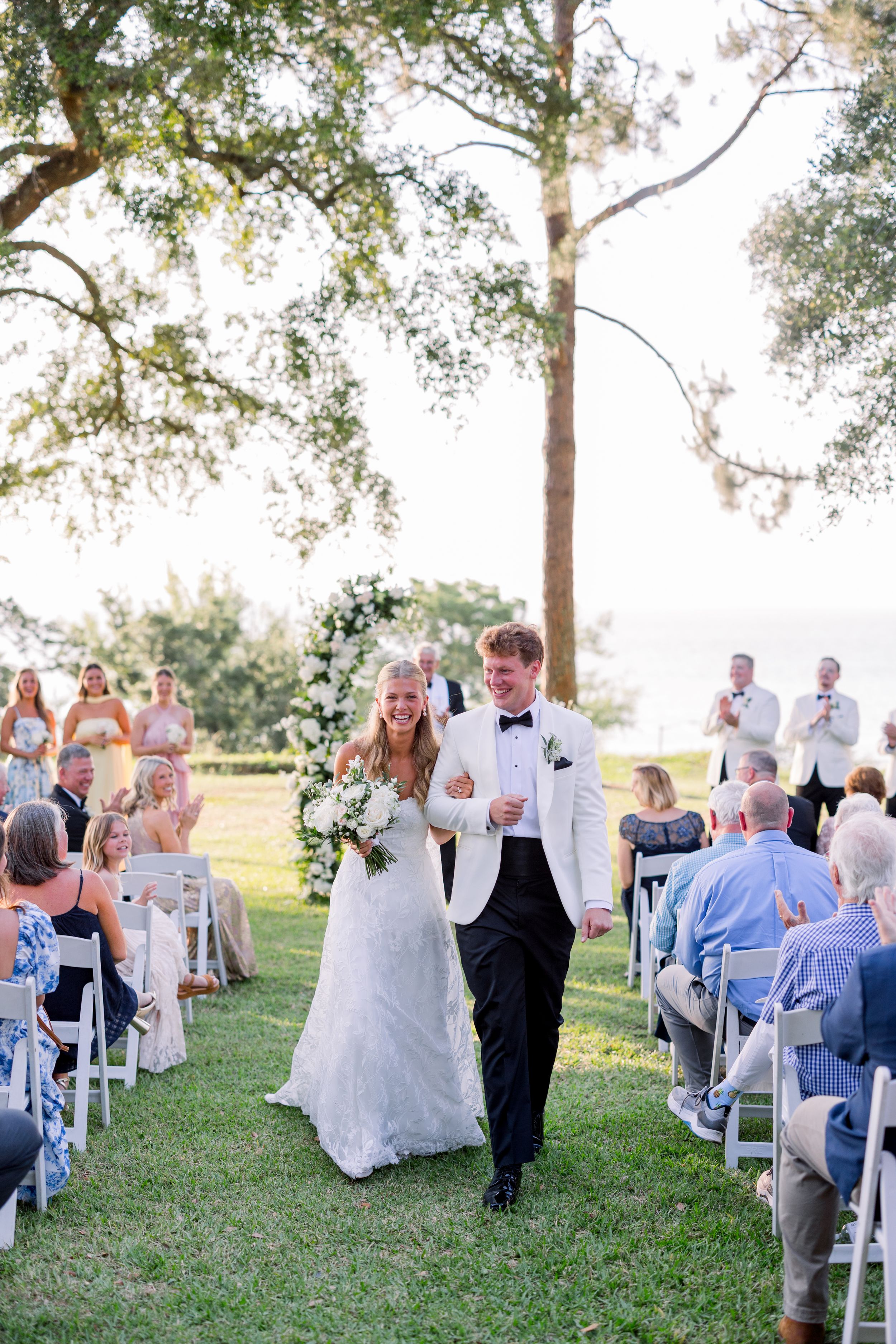 Wedding ceremony processional on a grassy lawn with white chairs and tall trees.