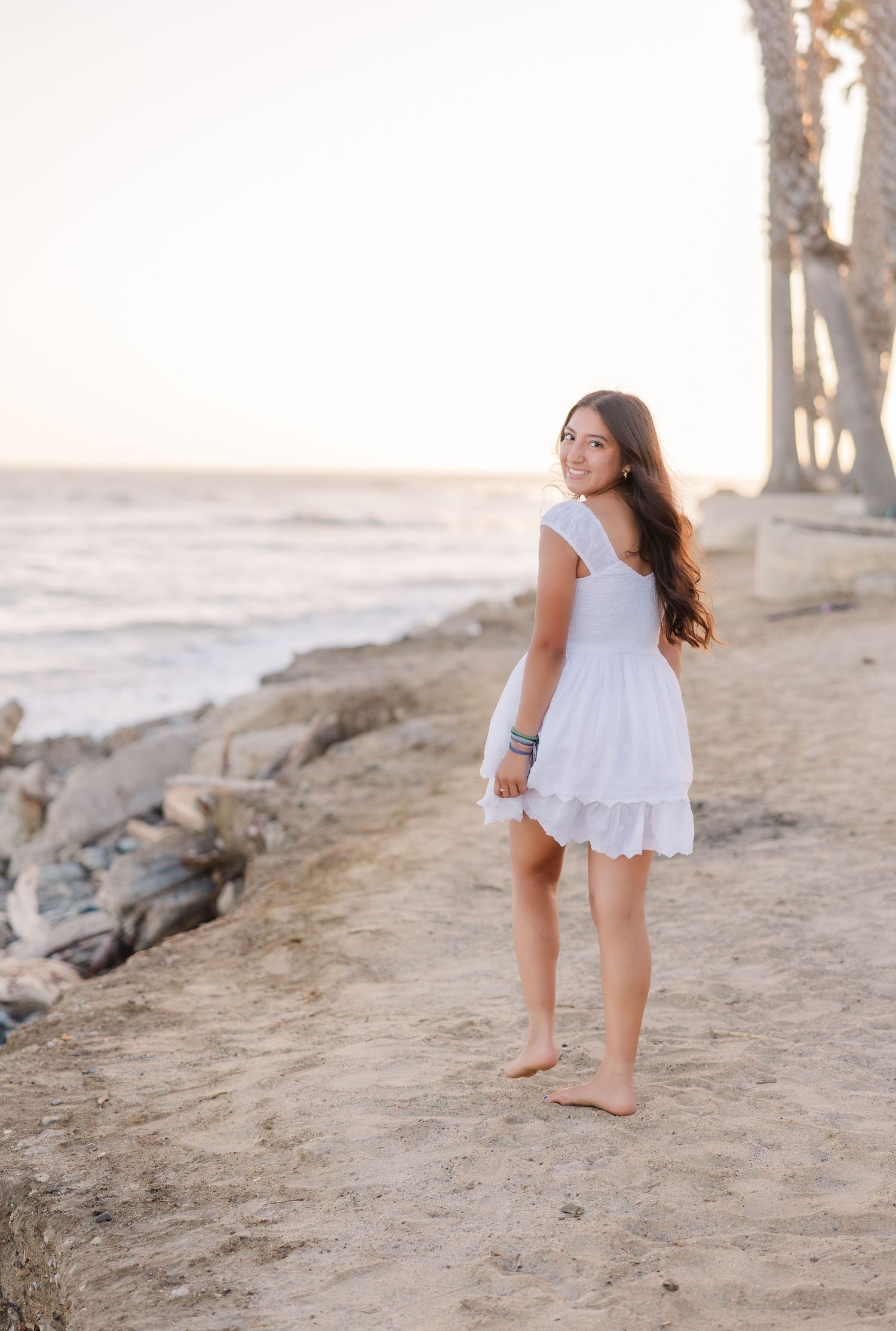A white eyelet dress flows in the ocean breeze as waves crash on a rocky beach at sunset.