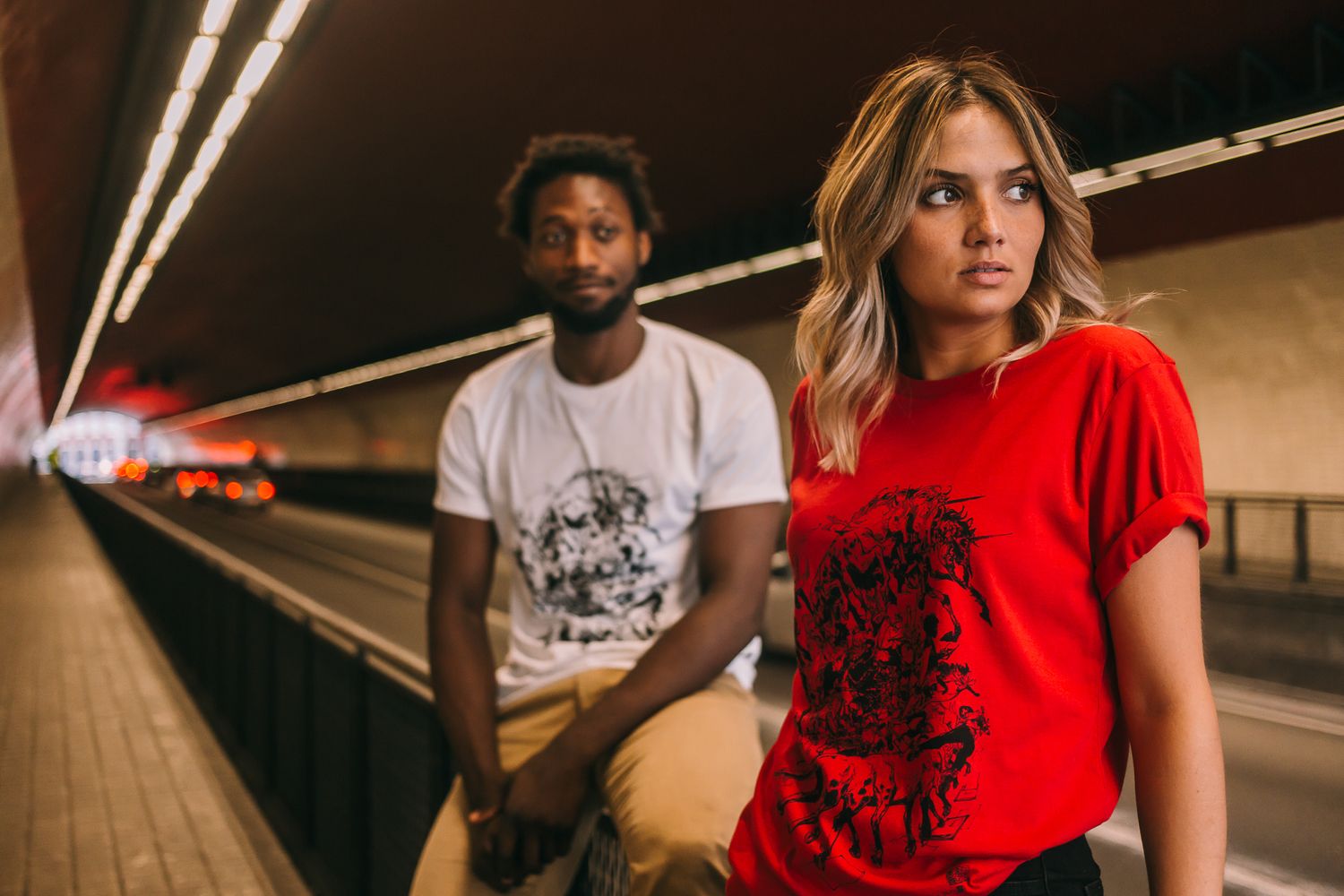 Two people in casual t-shirts pose in an empty subway tunnel with moody lighting and perspective leading lines.