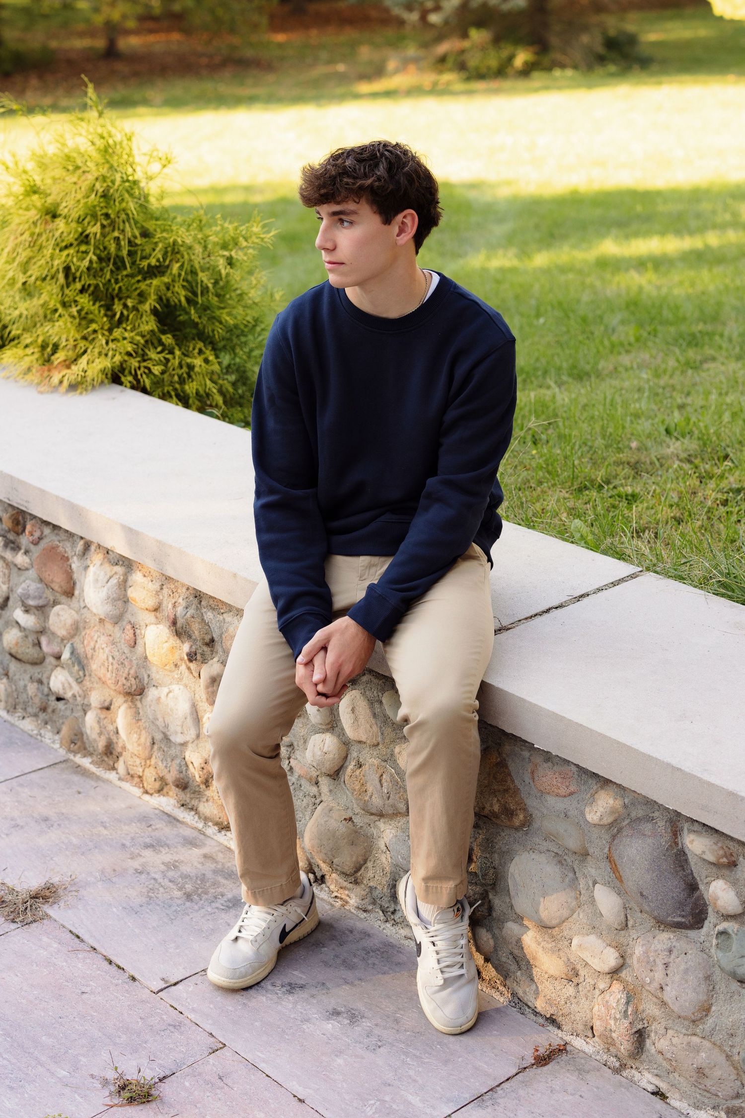 Person in a navy sweater and khakis sits on an outdoor stone wall on a sunny day.