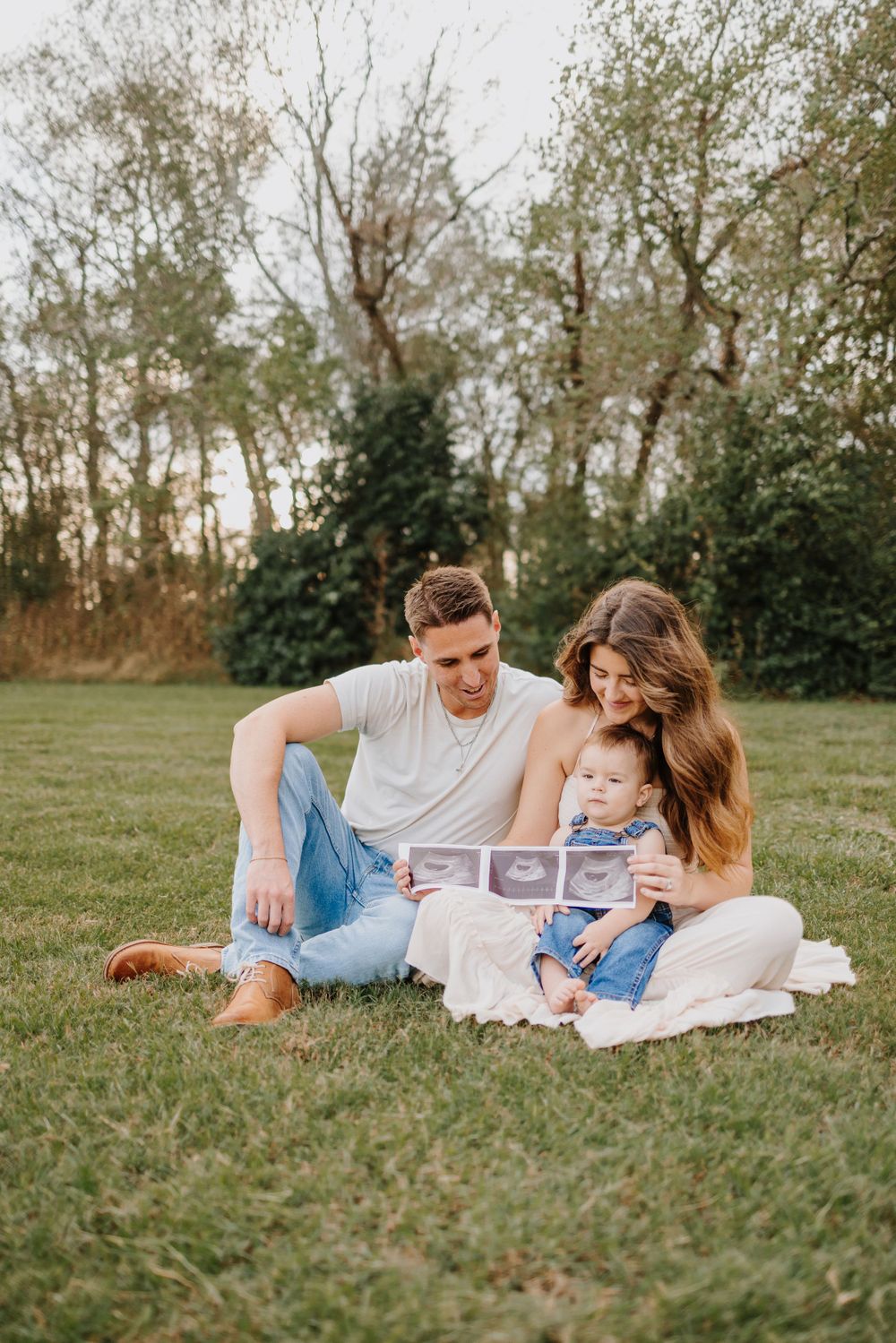 Family lounging on blanket in grassy field during sunset while looking at ultrasound photos.