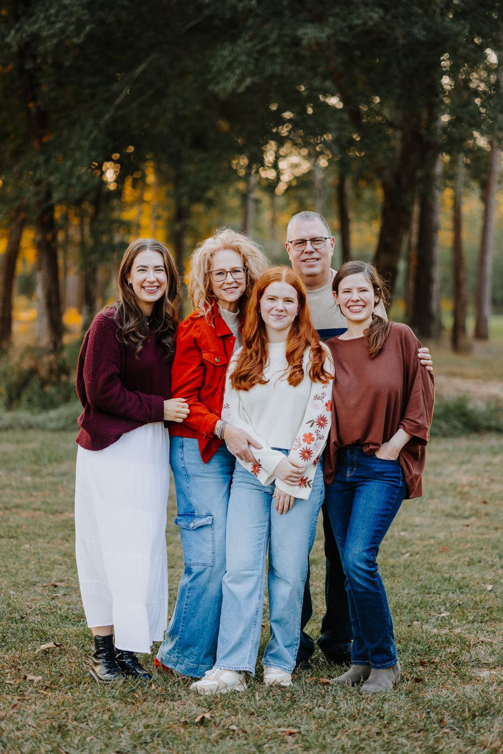 A group of four people wearing casual fall attire pose together outdoors in a wooded setting at sunset.