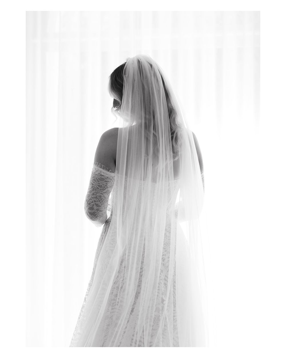 Black and white bridal portrait of a bride in a lace gown and flowing veil standing before a bright window.