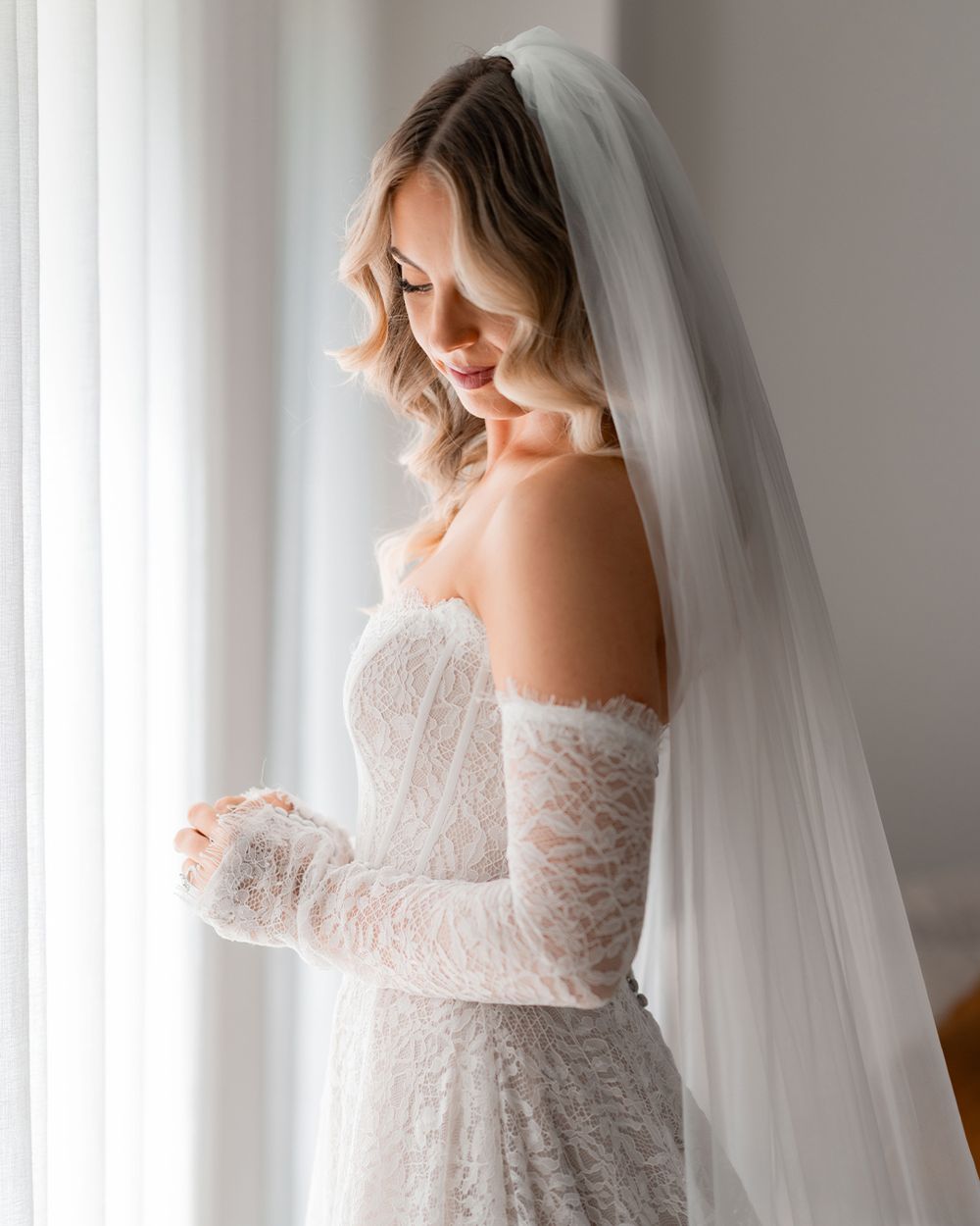 Bride in strapless lace wedding gown with long sleeves and white veil, gazing down near a bright window.