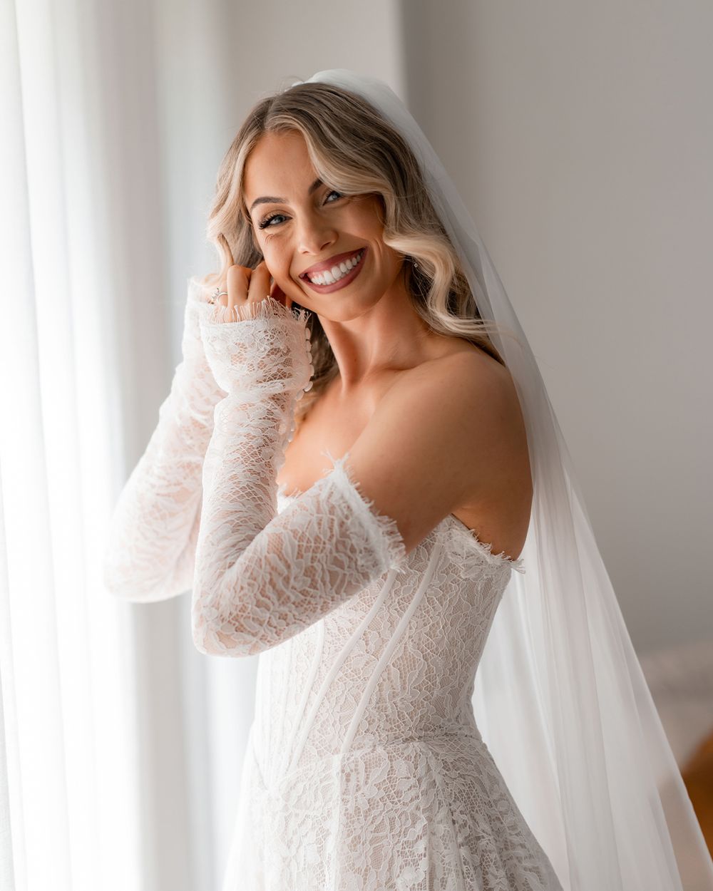 Smiling bride in off-shoulder lace wedding gown with long sleeves and veil, posing elegantly by a bright window.