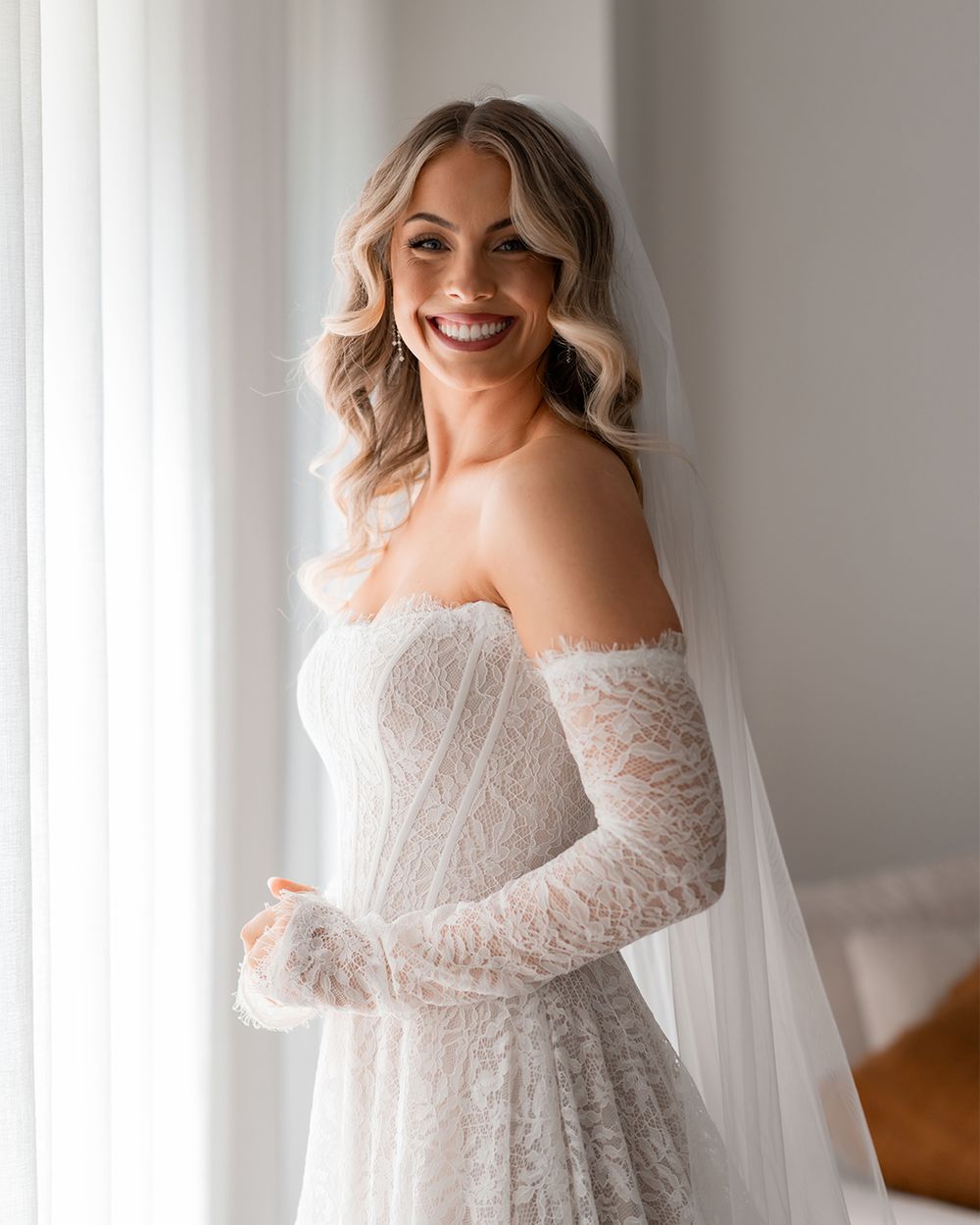 Smiling bride in strapless lace wedding gown with sheer sleeves and veil, posing elegantly near a bright window.