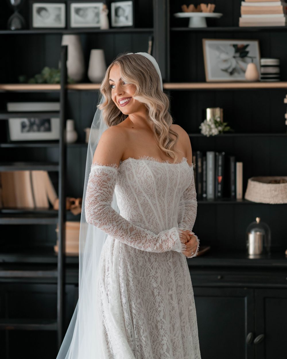 Smiling bride in off-shoulder lace wedding gown with veil, posing in front of dark bookshelves.