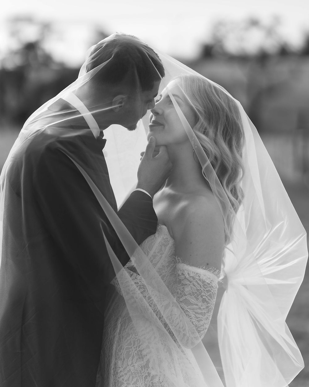 Black and white wedding photo of bride and groom sharing intimate moment under veil, touching foreheads romantically.