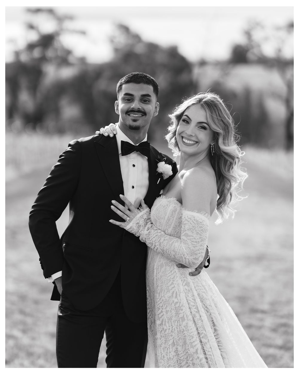 Black and white wedding photo of smiling bride in lace gown and groom in tuxedo posing outdoors.