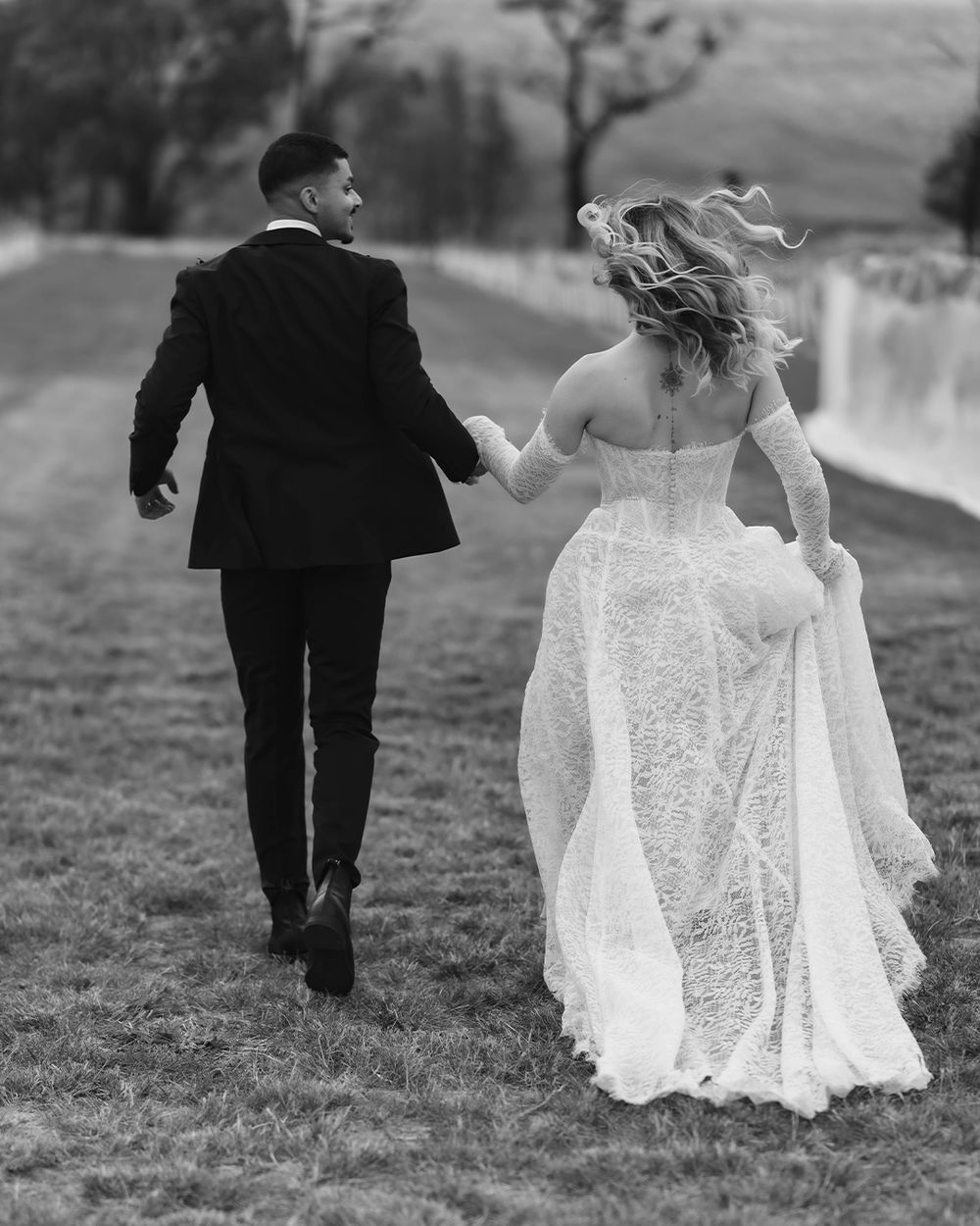 Black and white photo of bride and groom holding hands walking across green field, wedding dress flowing in wind.
