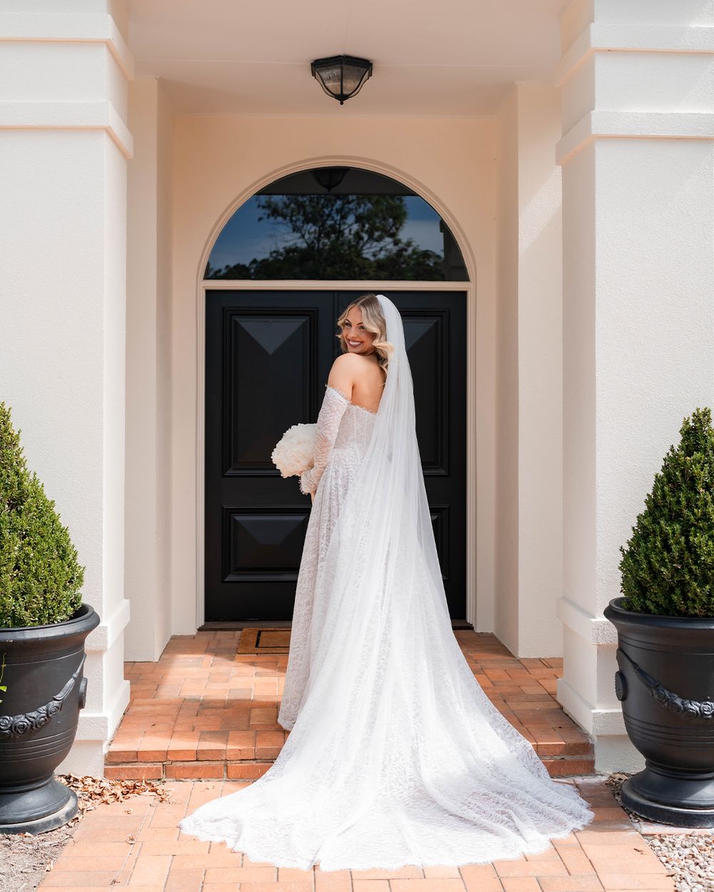 Bride in white lace gown and veil stands before elegant black arched door, holding bouquet, brick entrance with topiaries.