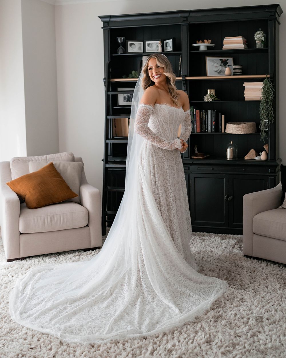 Bride in an off-shoulder lace wedding gown with a long train poses in an elegant room with dark bookshelves.
