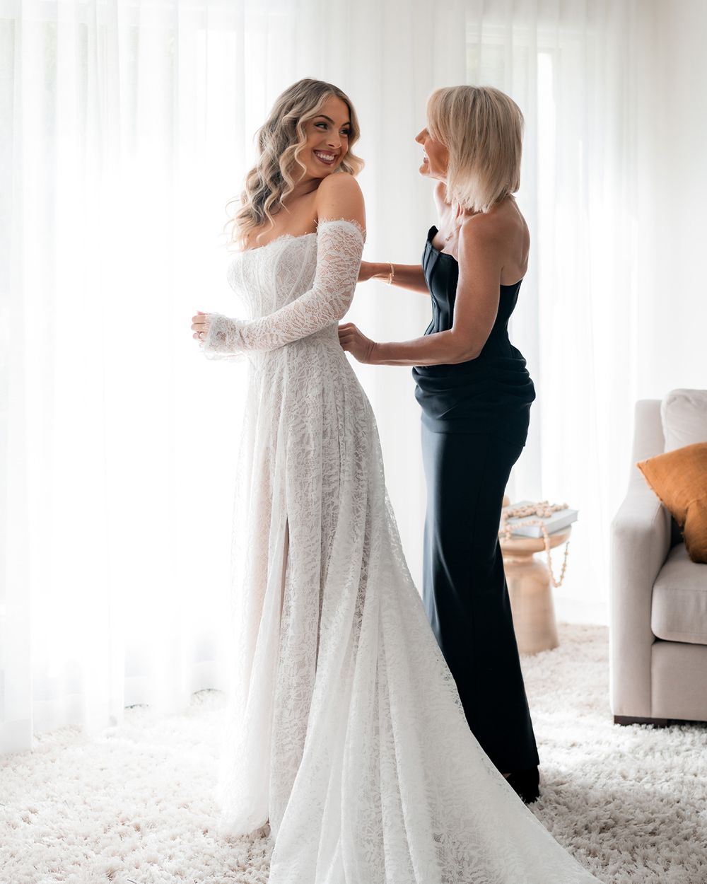 Bride in off-shoulder lace wedding gown being helped by bridesmaid in navy dress near bright window.