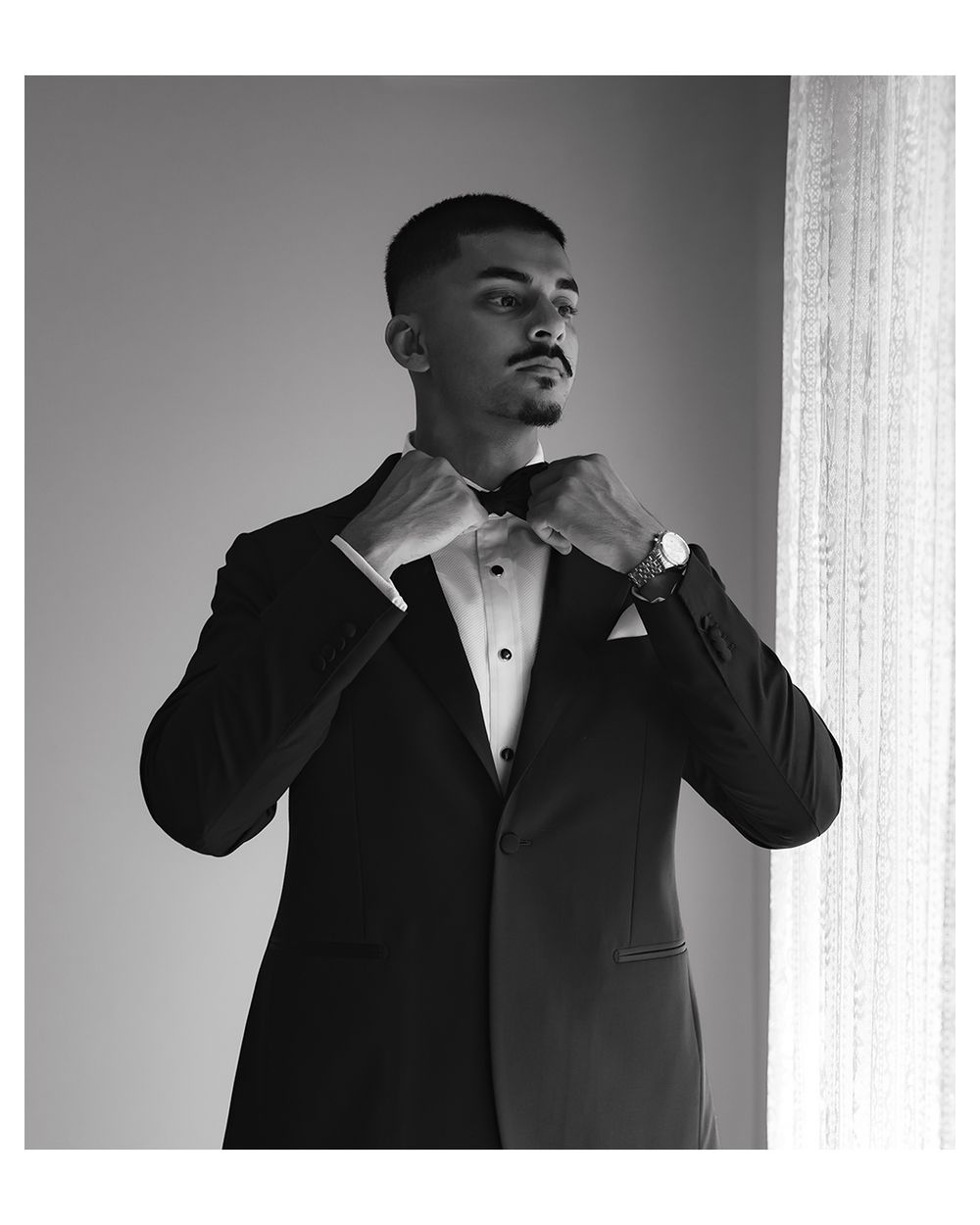 Black and white photo of a man in a tuxedo adjusting his bow tie, wearing a watch, standing near a curtained window.