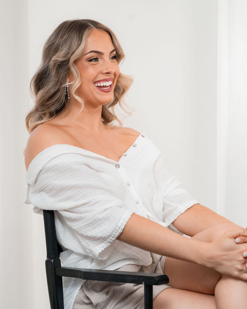 A smiling woman with wavy blonde hair sits in a chair wearing an off-shoulder white top and shorts, laughing joyfully.