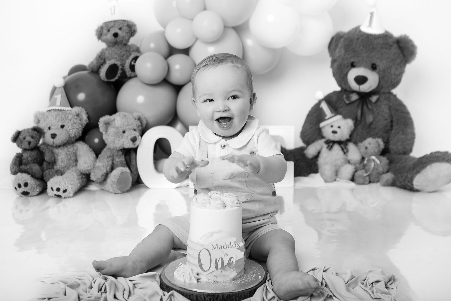 A black and white photo of a one-year birthday celebration with balloons and teddy bears arranged around the scene.