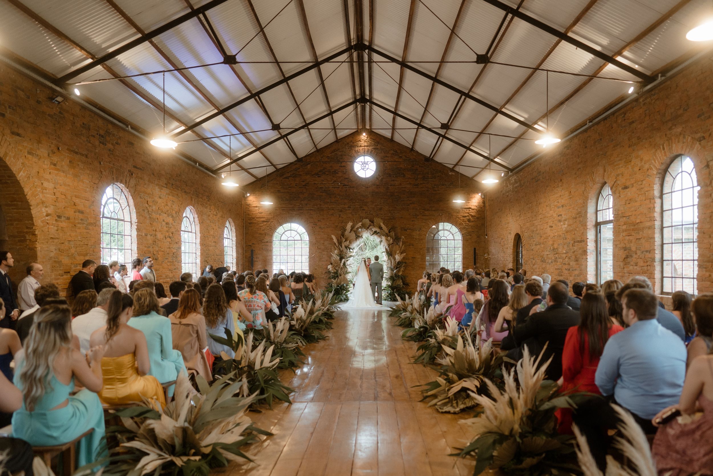Wedding ceremony in rustic chapel with exposed brick walls and rows of dried pampas grass aisle decorations.