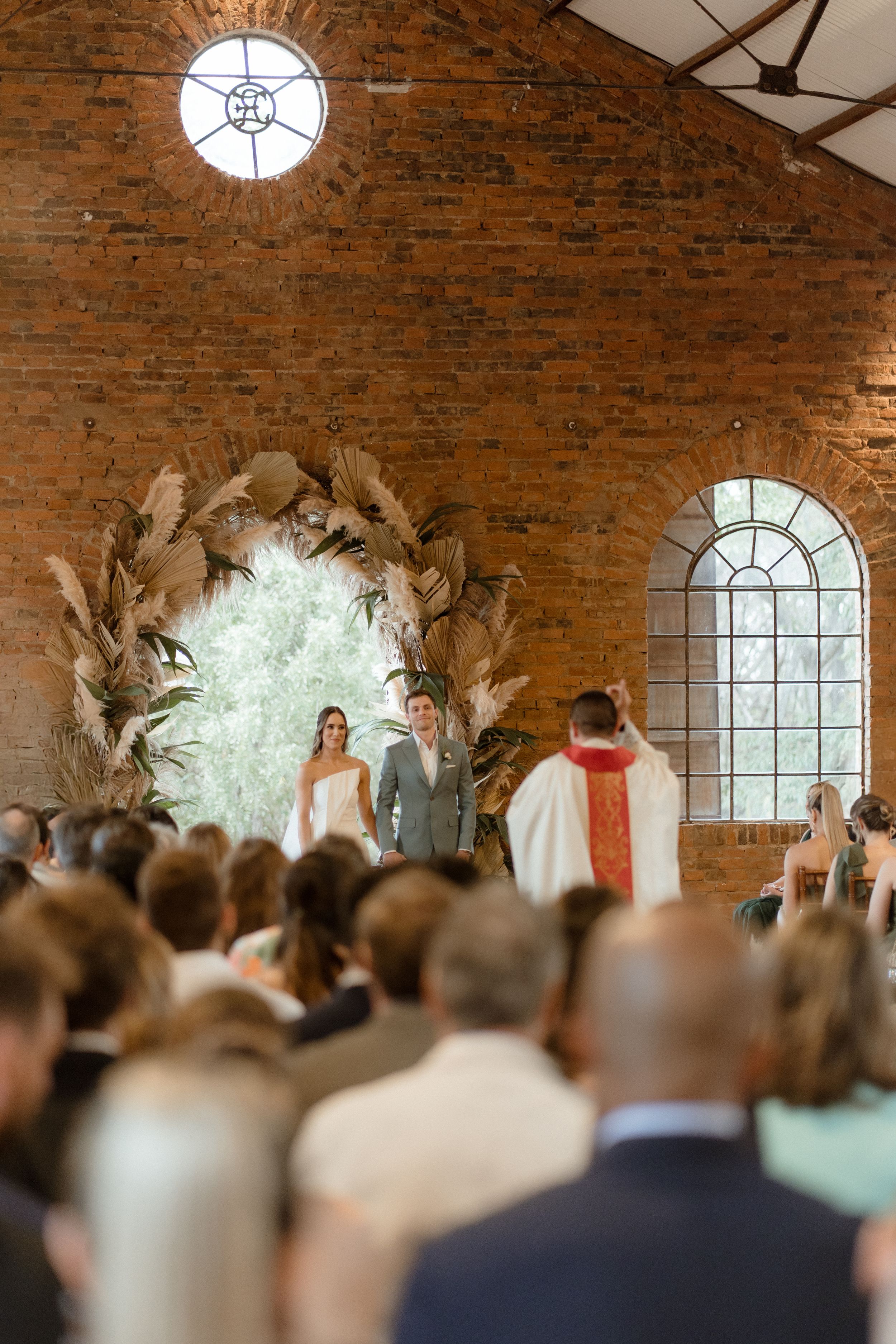 Wedding ceremony in brick chapel with large dried floral arch and arched windows providing natural light.
