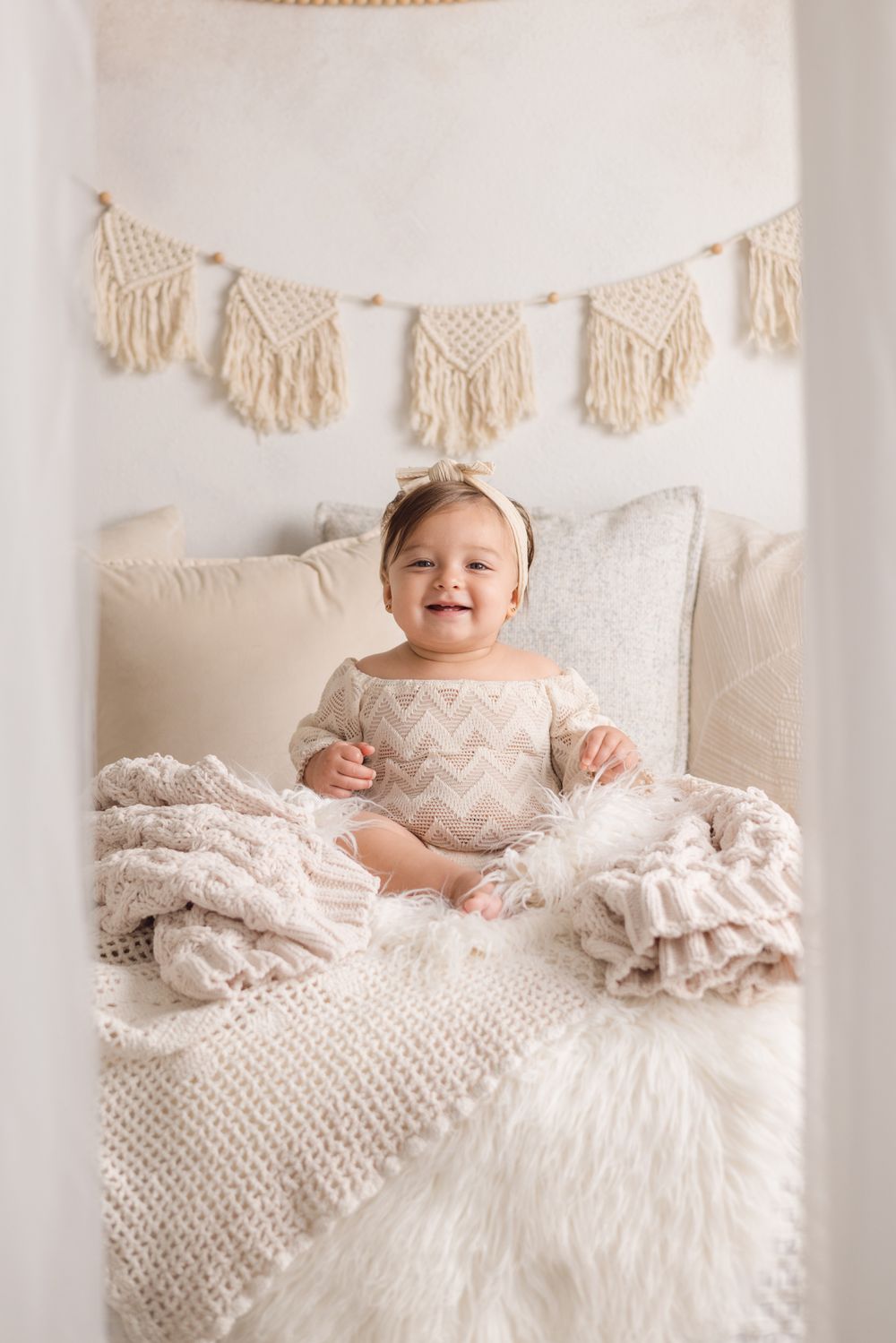Smiling baby girl in chevron romper sits on fluffy white fur rug surrounded by knit blankets in boho nursery.