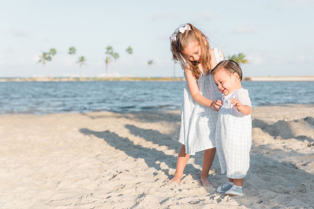 Two young children playing together on a sandy beach near calm water with palm trees in the background.