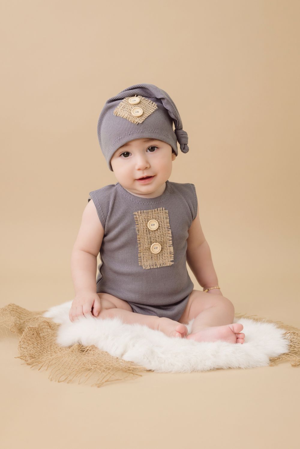 Baby in gray onesie and knotted hat with burlap button details, sitting on white fur rug against beige background.