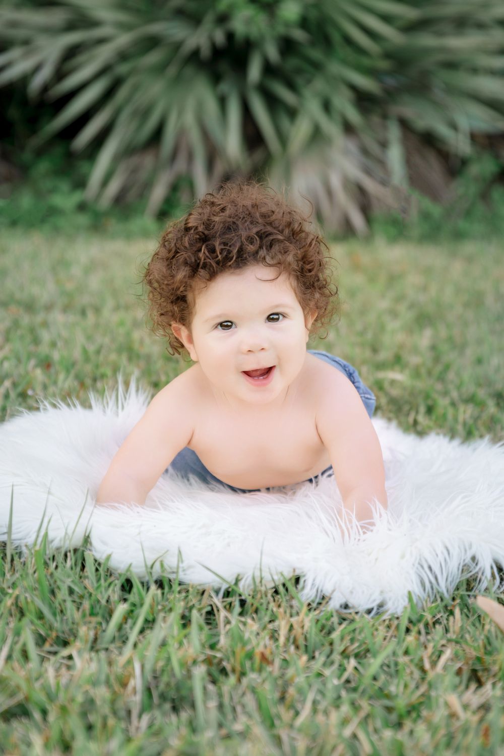 Curly-haired baby lying on white fluffy rug in grass, smiling outdoors with tropical plants in background.