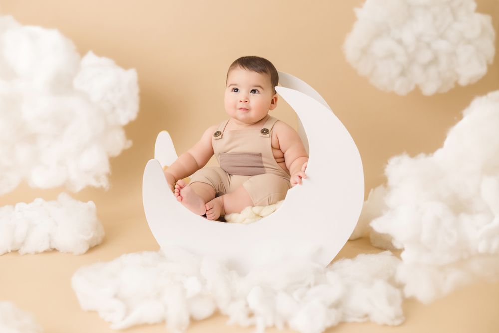 Baby in beige romper sitting on white moon prop surrounded by fluffy white clouds on warm beige background.