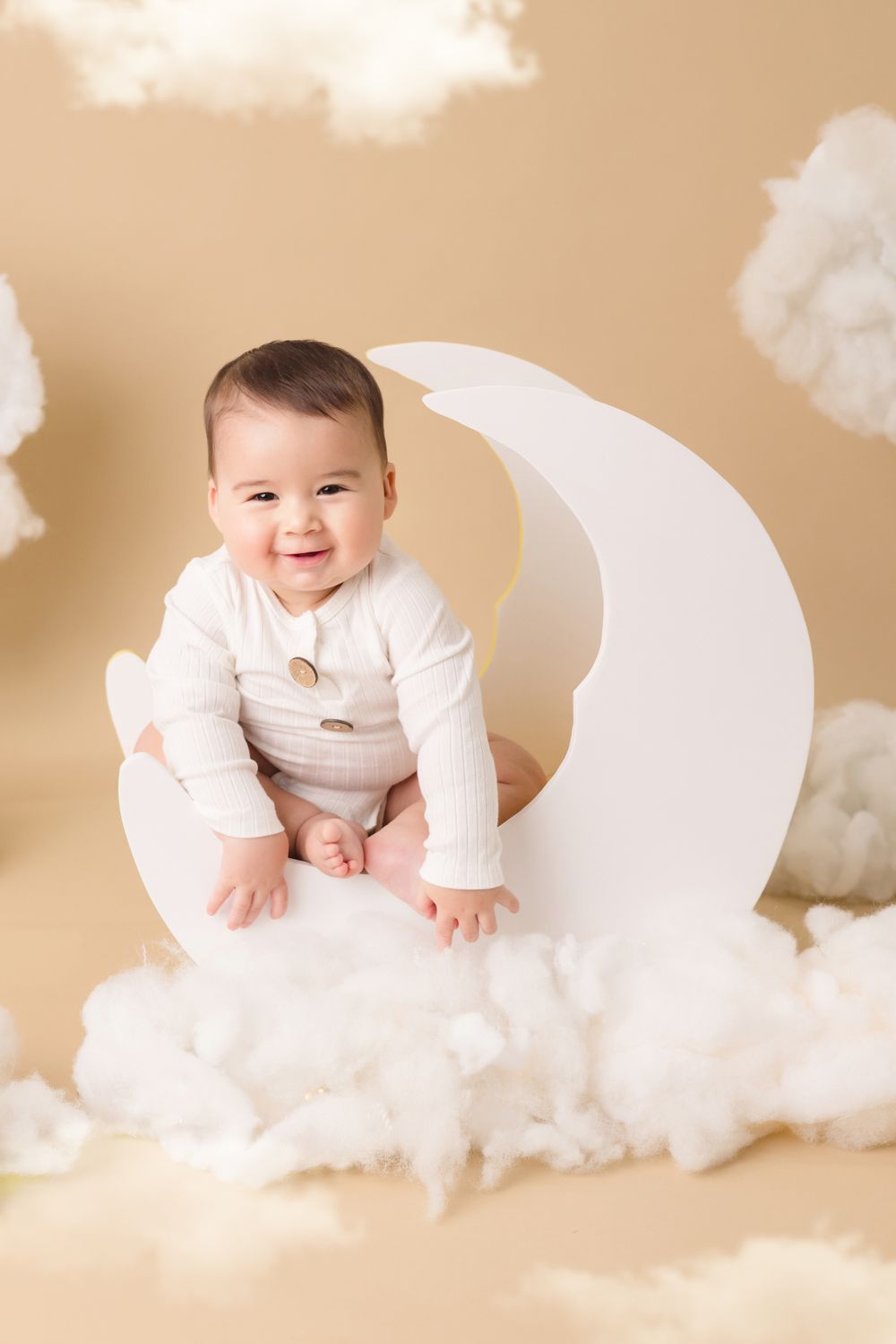 Smiling baby in white onesie sitting on a crescent moon prop surrounded by fluffy clouds on a beige background.