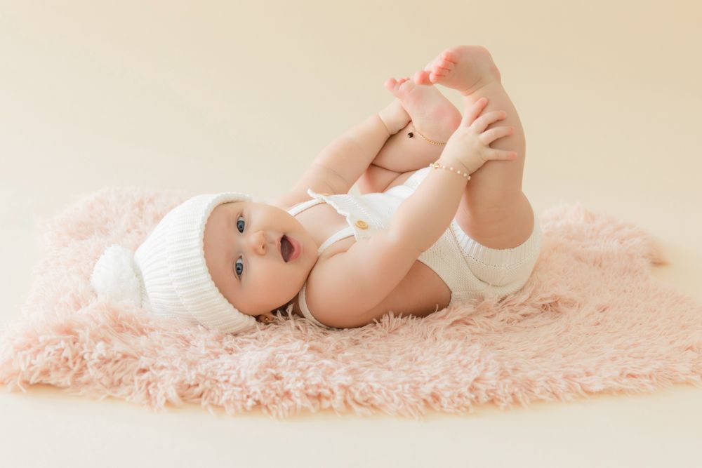 Adorable baby in white bonnet and bodysuit lying on pink fluffy rug, playfully holding feet up in the air.