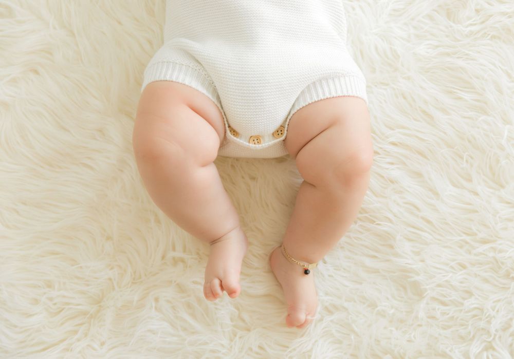 Chubby baby legs resting on a soft white fluffy rug, wearing a white onesie with a delicate gold anklet.
