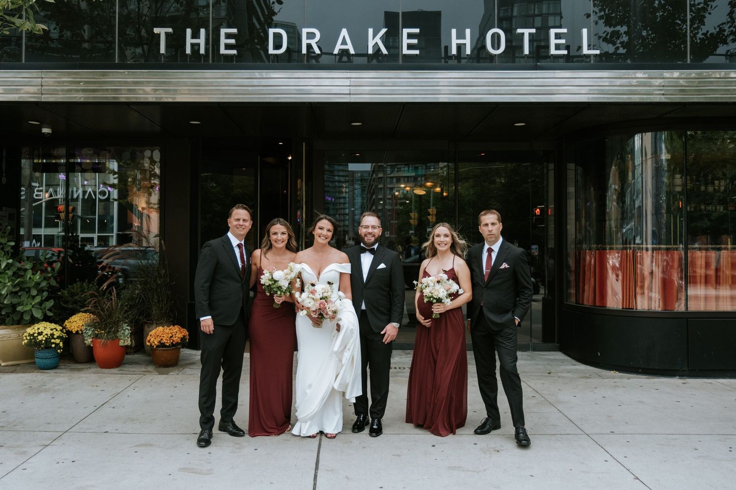 Wedding party in burgundy and black formal attire poses outside The Drake Hotel entrance.