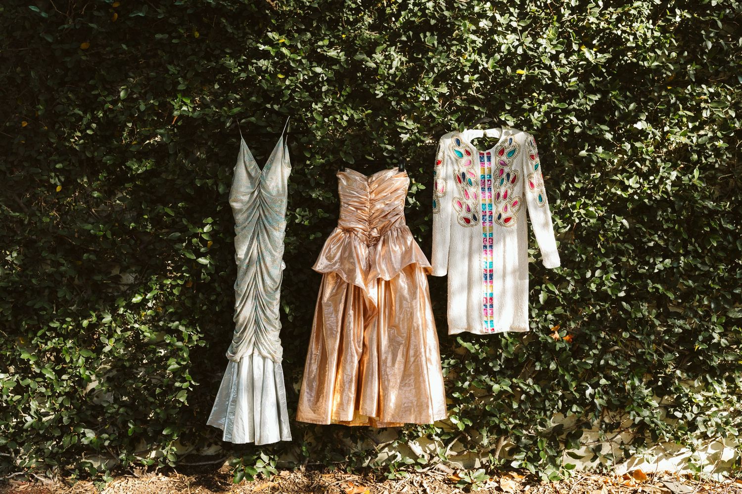 Three vintage dresses hanging outdoors against green foliage, featuring floral embroidery and feminine designs.