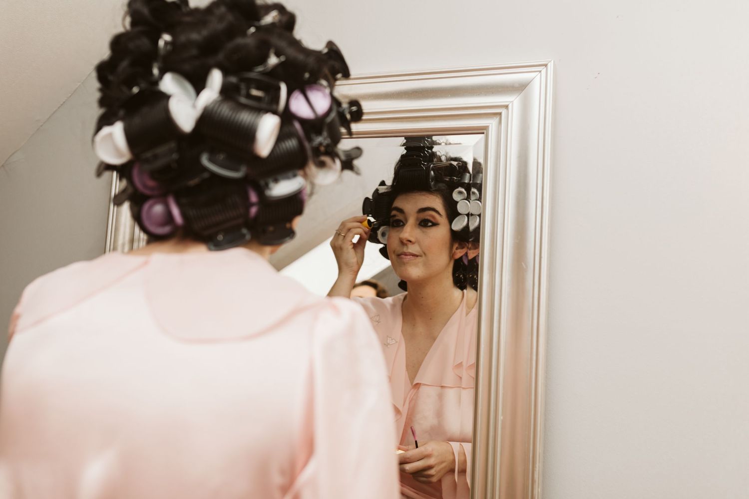 Woman in a pink robe getting ready with hair curlers in a brightly lit room.
