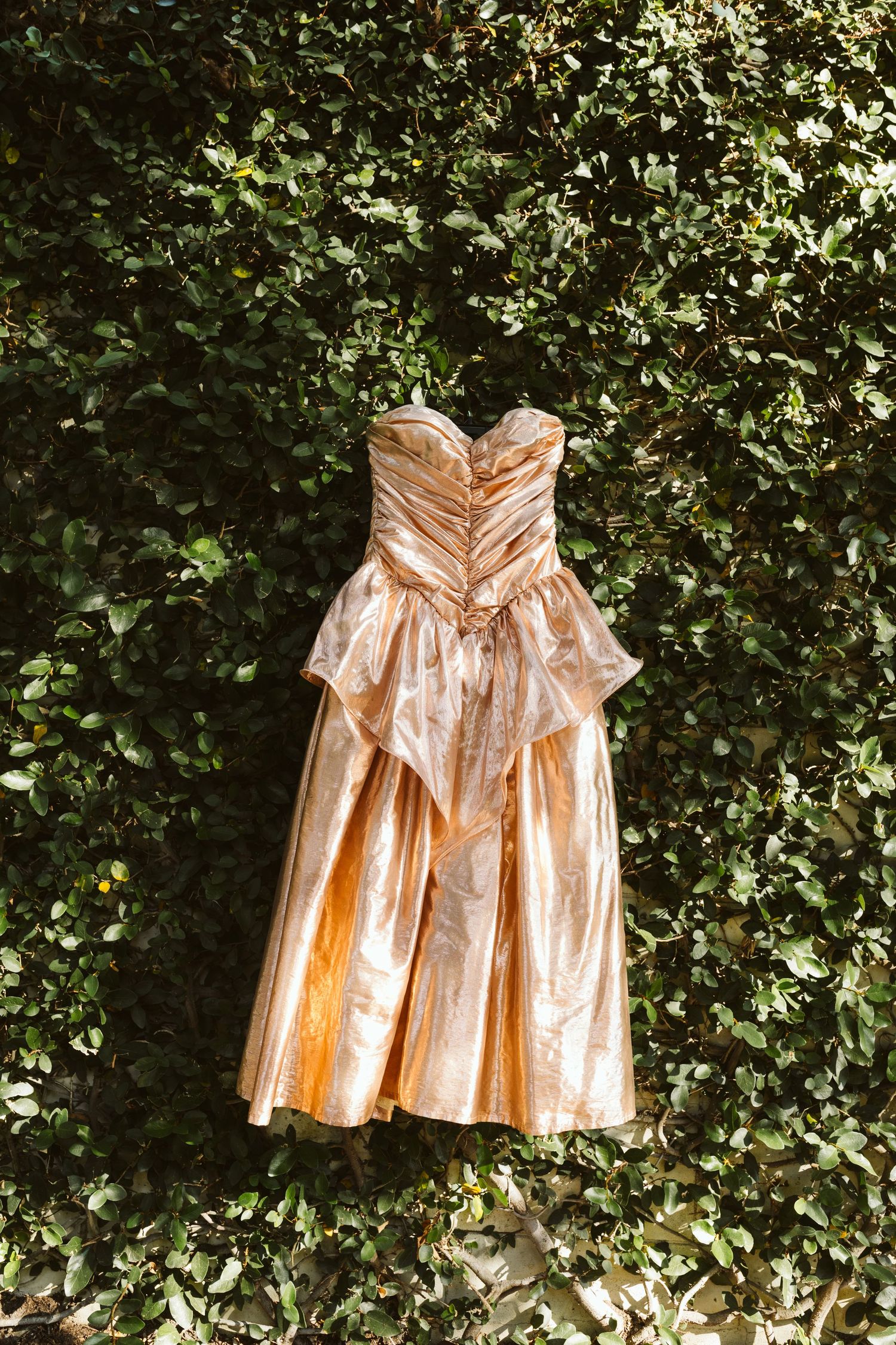 Gold metallic dress displayed against a lush green hedge wall backdrop.