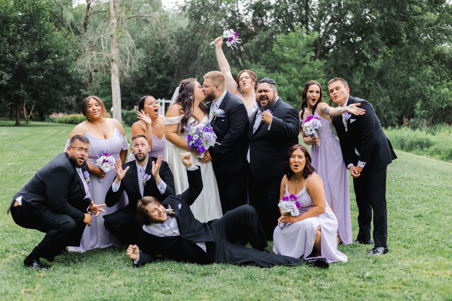 Wedding party striking fun poses on grass in formal attire with bridesmaids in purple dresses.