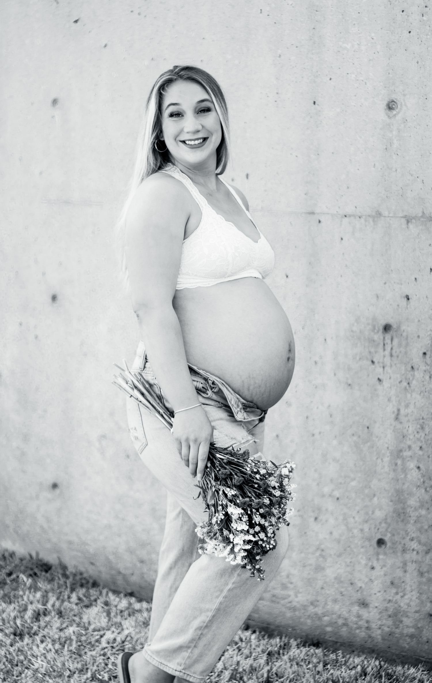 Black and white maternity photo showing a pregnant woman in casual attire holding dried flowers against a concrete wall.