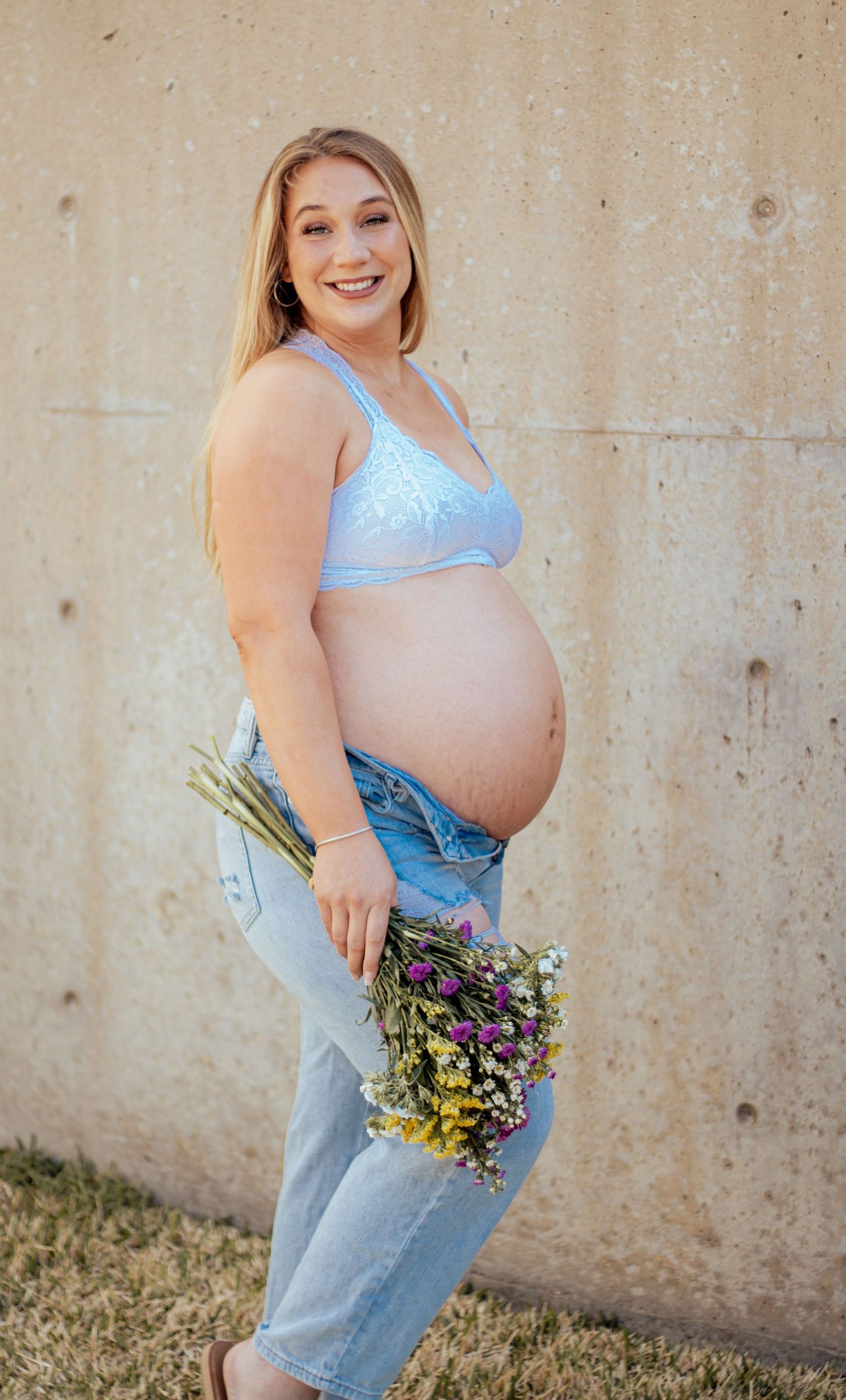 Maternity photo showing pregnant person in distressed jeans and blue bra top holding wildflowers against beige wall.
