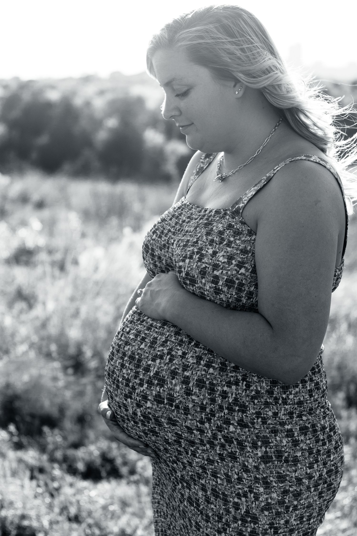 Black and white portrait of an expectant mother in a floral sundress standing outdoors in a sunlit meadow.
