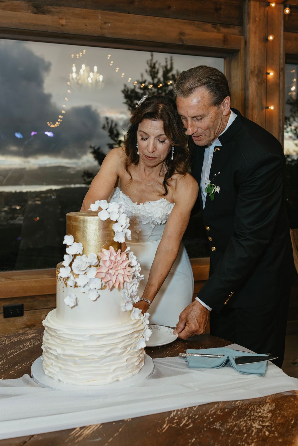 A wedding couple cuts into their elegant white tiered cake decorated with orchids at a rustic reception venue.