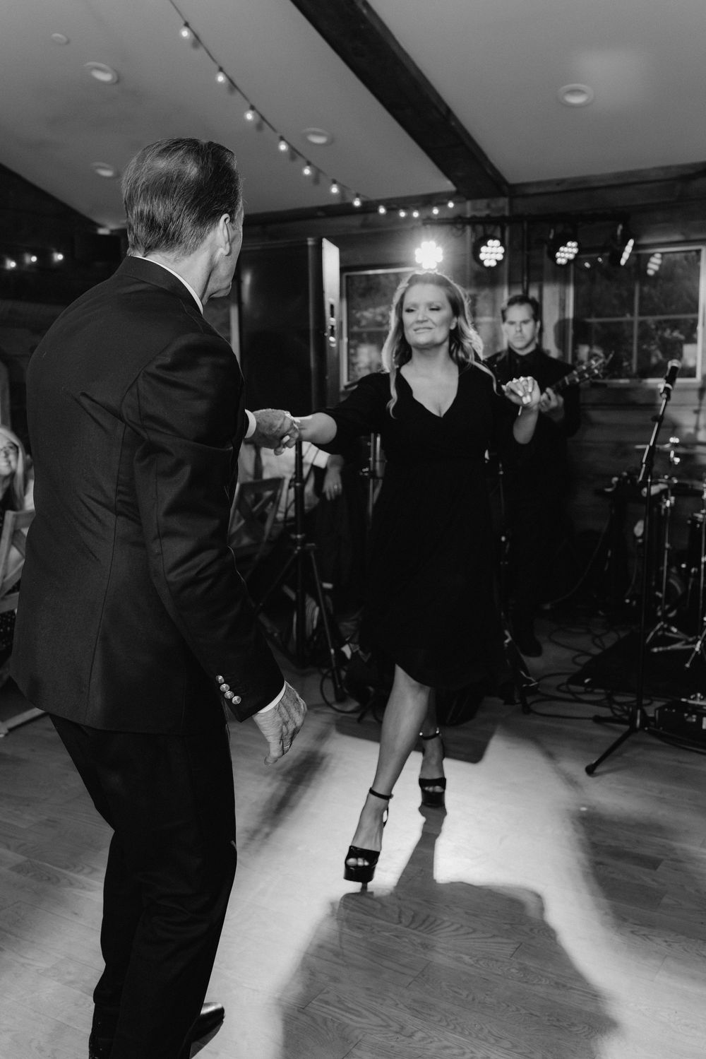 Black and white photos of wedding guests dancing on a hardwood floor under string lights at an indoor reception.