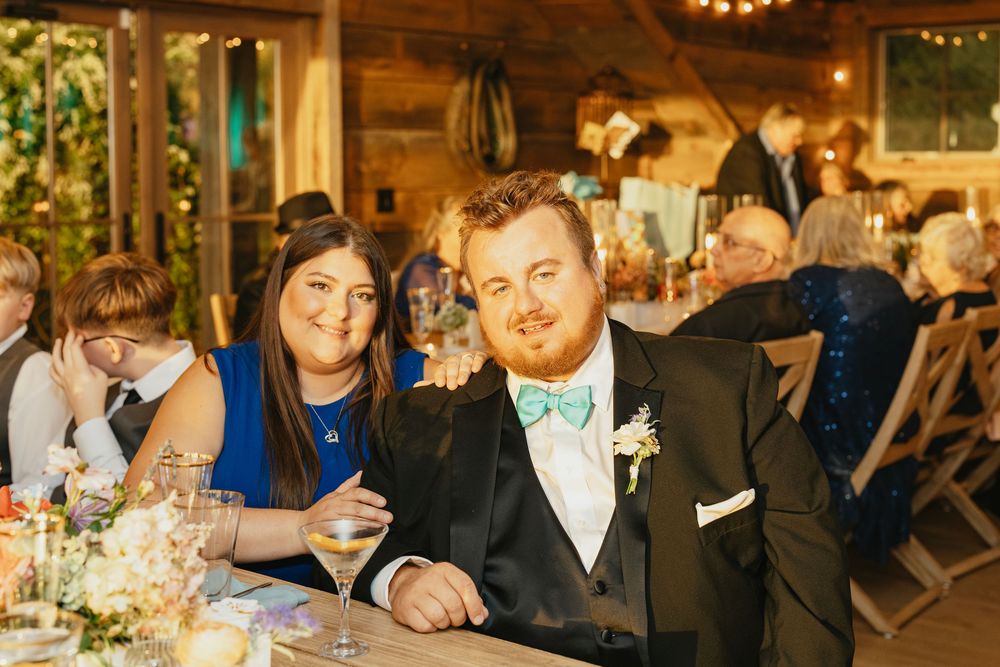 Wedding guests gather around tables at an indoor reception venue with warm lighting and elegant decor.