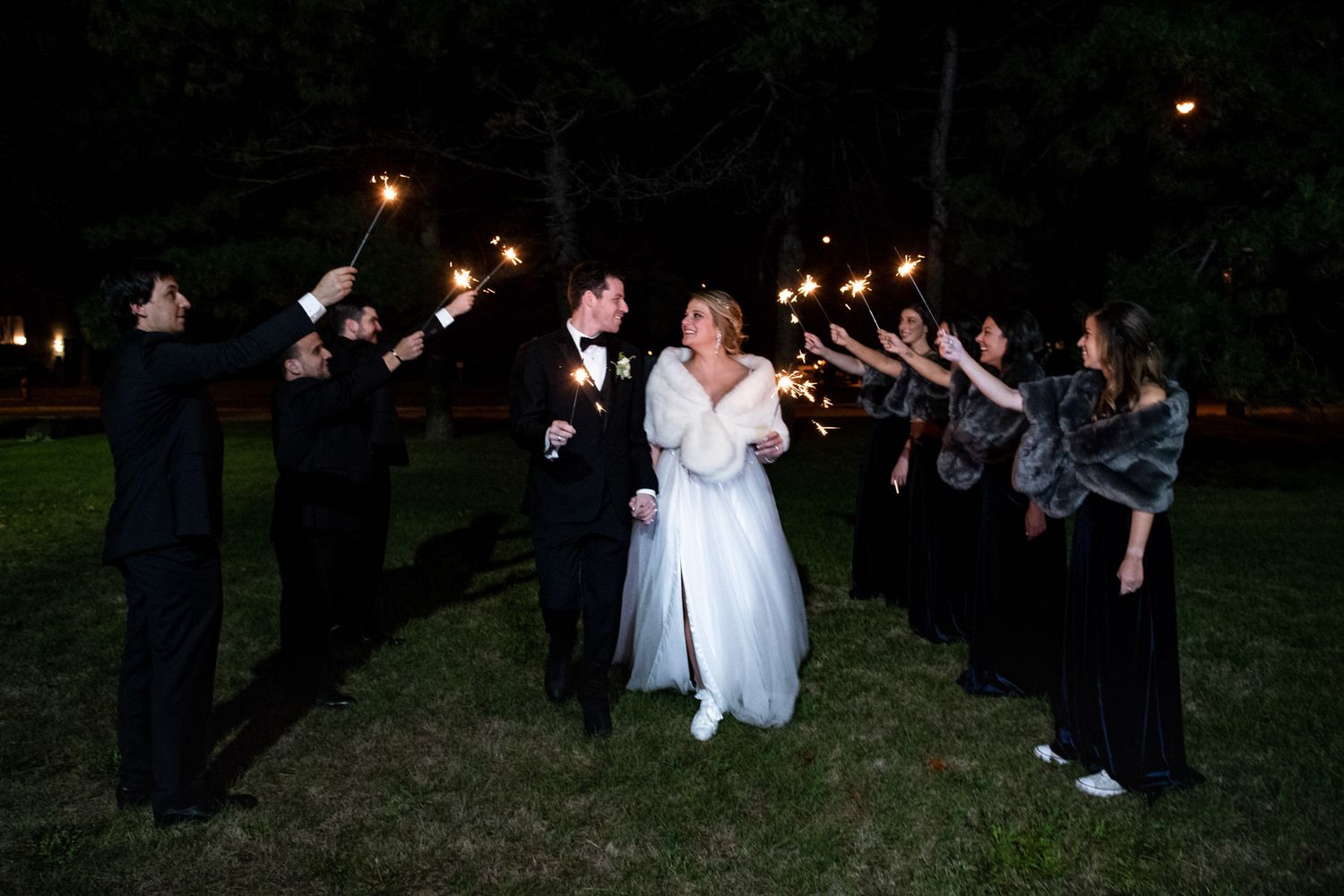 Wedding couple walks through sparkler tunnel with bridal party at outdoor nighttime reception.