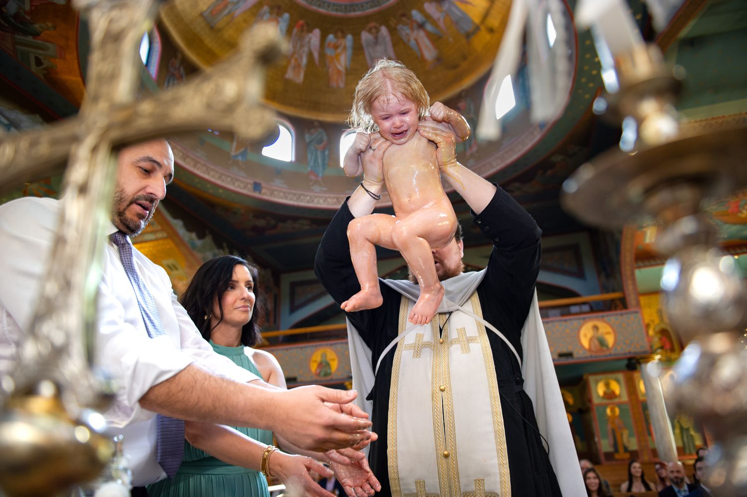 Orthodox priest baptizes crying baby over font as Godparents watch at St. Demetrios Orthodox Church in Toronto