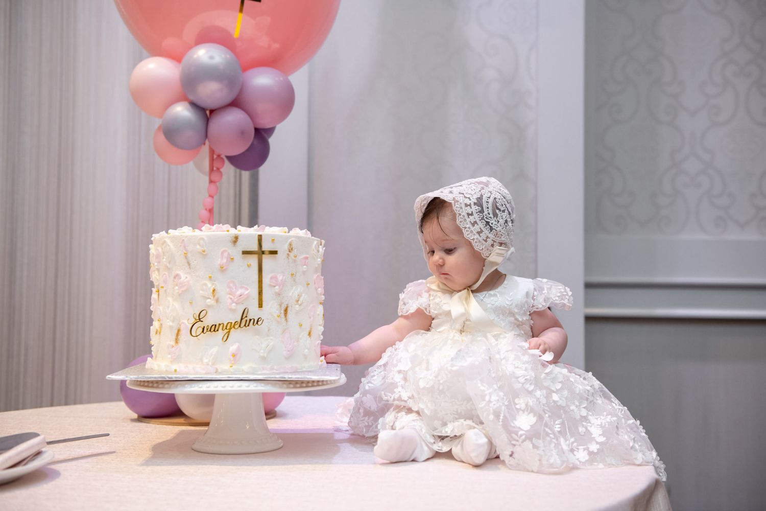 Baby girl in white christening gown and bonnet touches decorated baptism cake with cross.