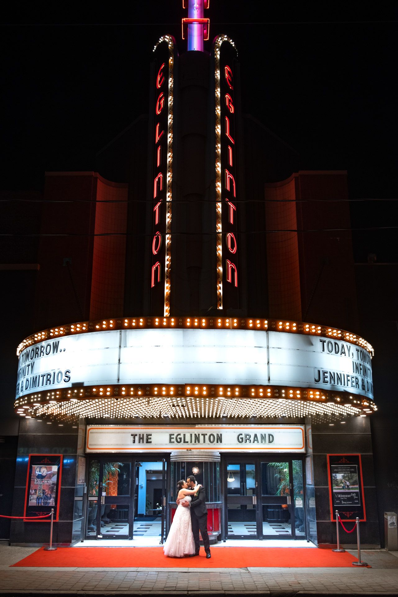 Bride and groom embrace on red carpet outside illuminated Eglinton Grand theater at night.